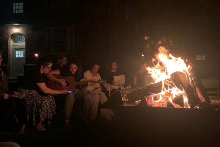 Students gather around the bonfire while some sing and one plays guitar