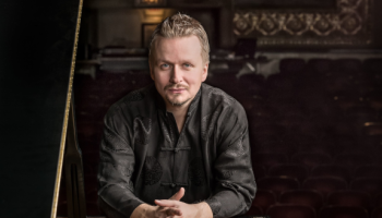 A man in an ornate room looking at the camera with his hands folded.