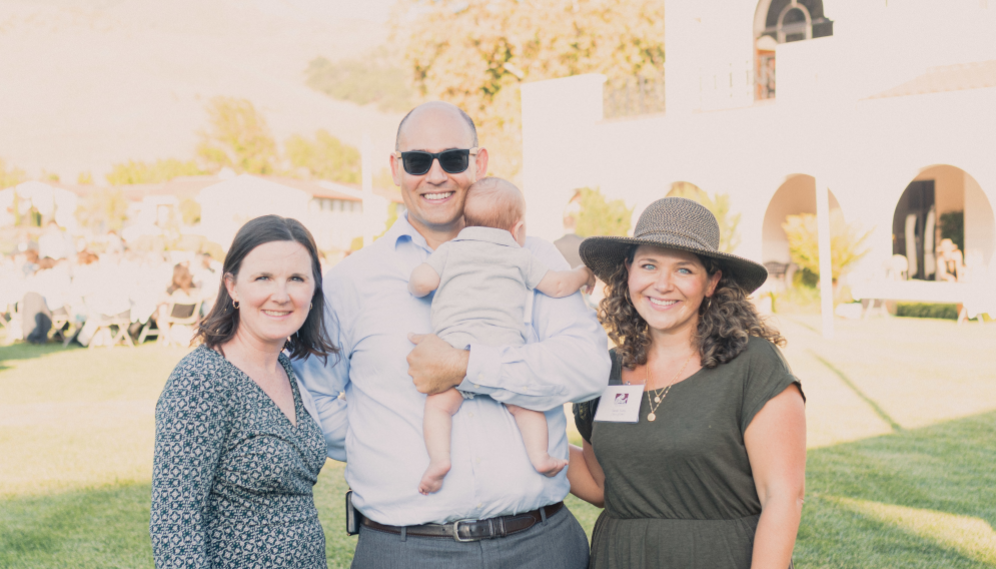 Three pose for a photo, the man in the middle with a baby in hand