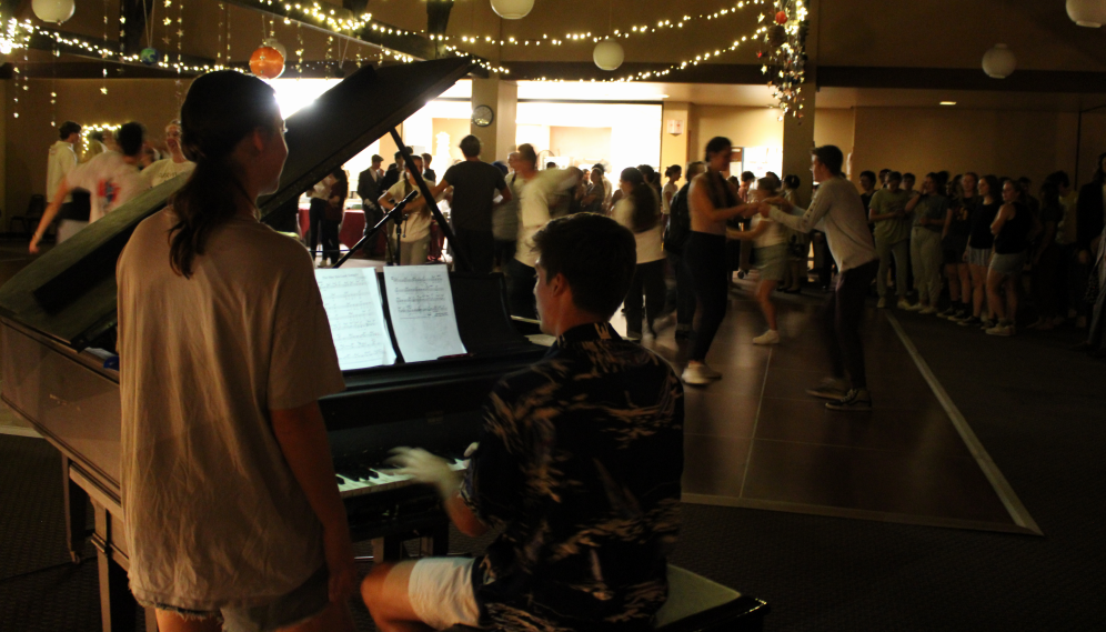 A pianist performs, while a page-turner stands beside him, and the students dance in the background