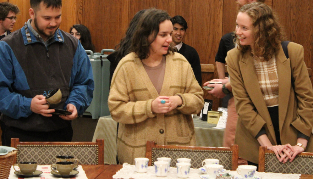 Behind the teacups neatly laid out, two chat and another selects a cup