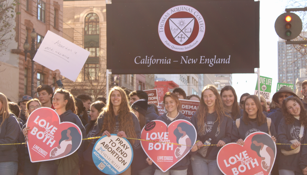 Students holding signs reading "Love them Both" march in front of the TAC banner