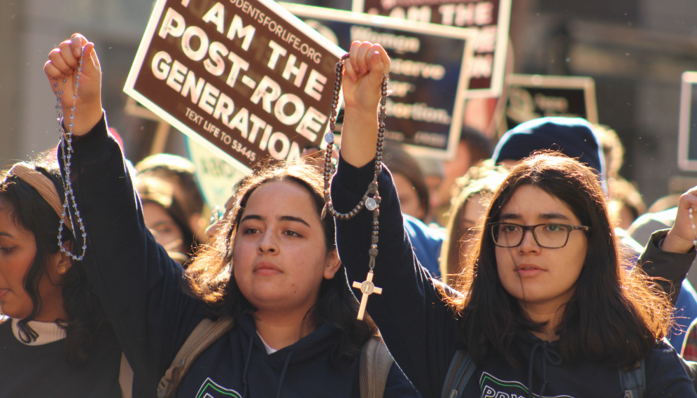 Two hold up Rosaries in front of a sign reading "I am the Post-Roe Generation"