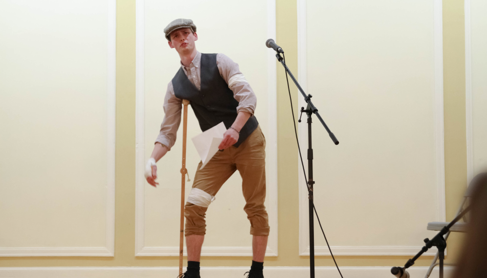 A student performs in flatcap, faux knee-breeches, and a crutch
