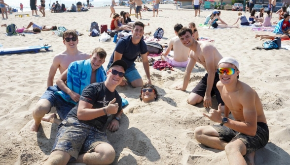 Six "thumbs-up" around their comrade, who is buried up to the neck in sand and grinning
