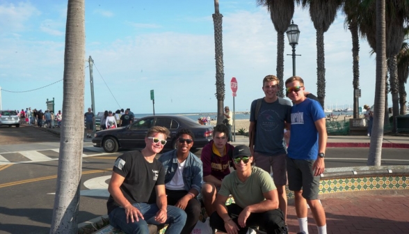 As the sun begins to set, six guys pose at a low tiled bench afront a street and the beach