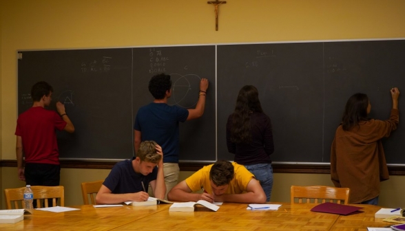 Four students practicing at the board, with two others studying at the table behind them