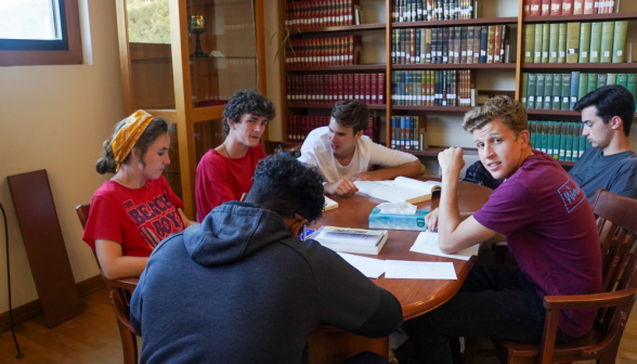 Six students at an oval table afront neatly organized bookshelves