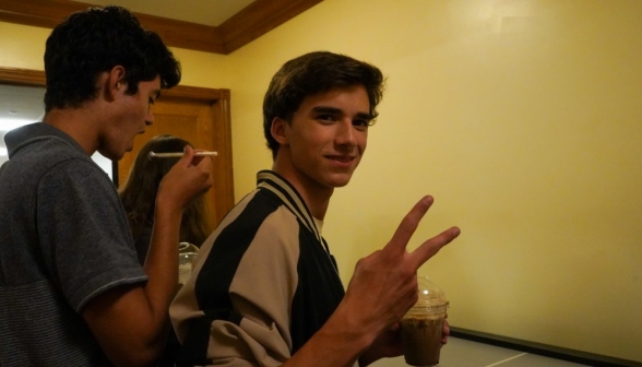 In the foreground, a student makes the peace sign with a coffee in the other hand. In the background, one is attempting to eat whipped cream with their straw