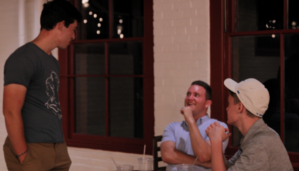 A standing student chats with John Jost and another student, who are seated at an upper-level table afront a window