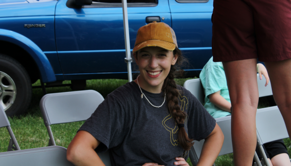 A student seated in a row of chairs, wearing a baseball cap and smiling at the camera