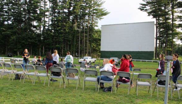 Students begin to fill in the rows of chairs outside on the grass at the drive-in