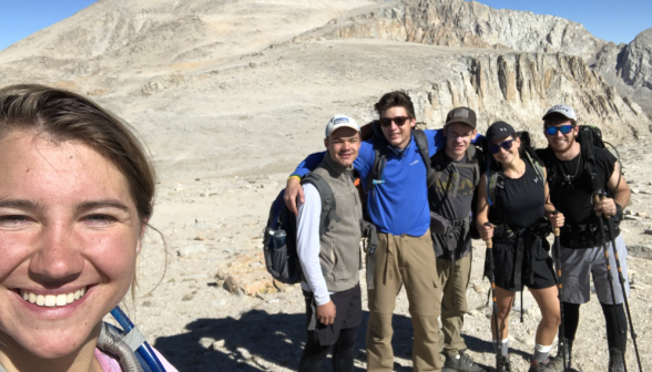 One student takes a picture, with five others posing in the background with their hiking equipment