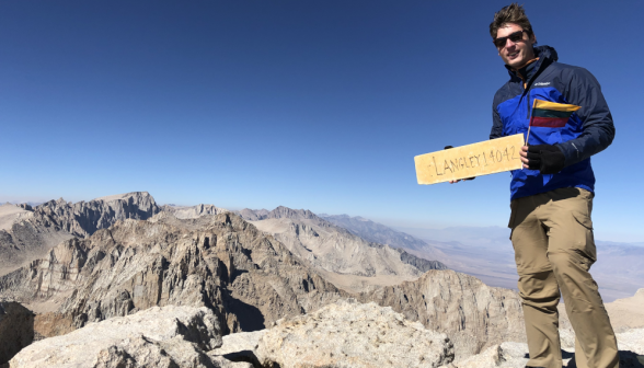 Another student holds up the "Langley 14042" sign, standing on a rock at the edge of a steep valley edge of a 