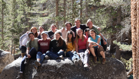 Thirteen students pose for a picture, seated atop a large boulder