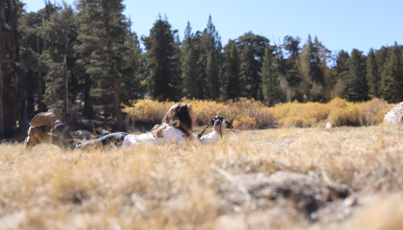 A student lies full-length in the long dry grass with her camera to snap an image