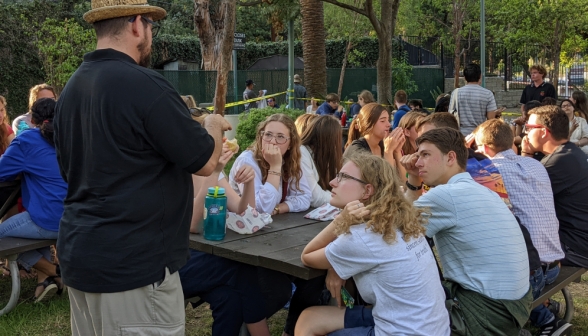 Students at a picnic table outside give their attention to a man in a woven fedora