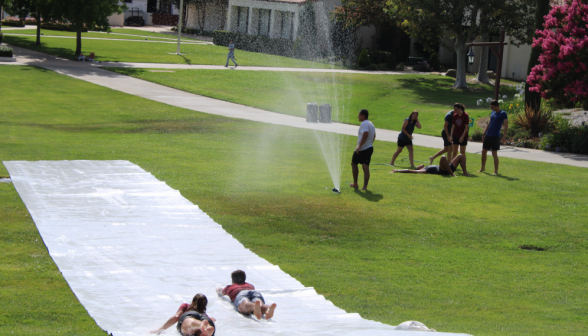 Long shot: students go down the slide and jump around in a nearby sprinkler. One relaxes, flopped on the ground