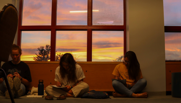 Three sit cross-legged on the floor, studying. Behind them, a glorious sunset through the window