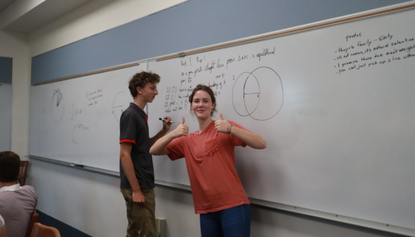 A student gives "thumbs-up" while another practices a prop at the board behind her