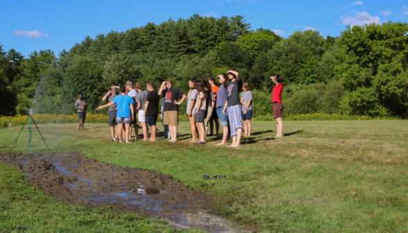 The freshmen in a group on the field, behind a sprinkler moistening the mud pit