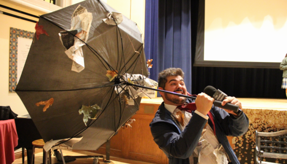 A student dressed as a weather reporter in an intense wind, with an inverted umbrella