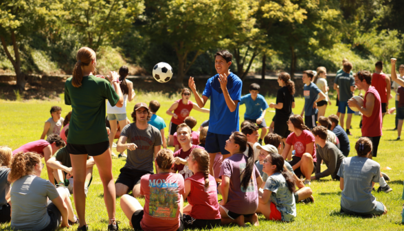 Many sit on the grass while two pass a ball