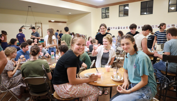 Long shot of a crowded coffee shop, with two at a table smiling for the camera in the foreground