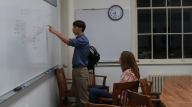 A student practices demonstrating props at the board to another student