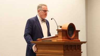 A shot from the left of a man standing behind a podium looking out ahead of himself.